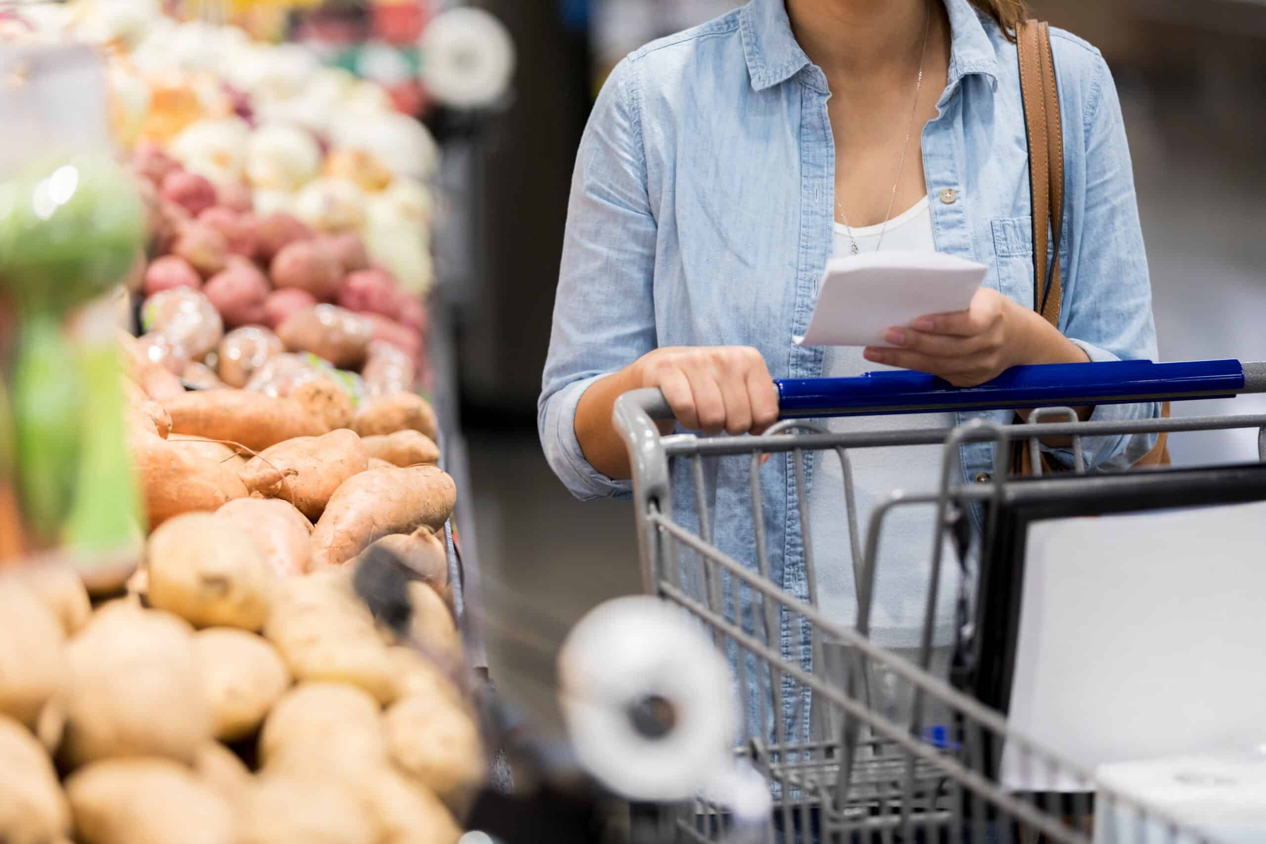 A Woman Pushes Her Cart Past The Potatoes In The Grocery Store.  She Holds A Shopping List.
