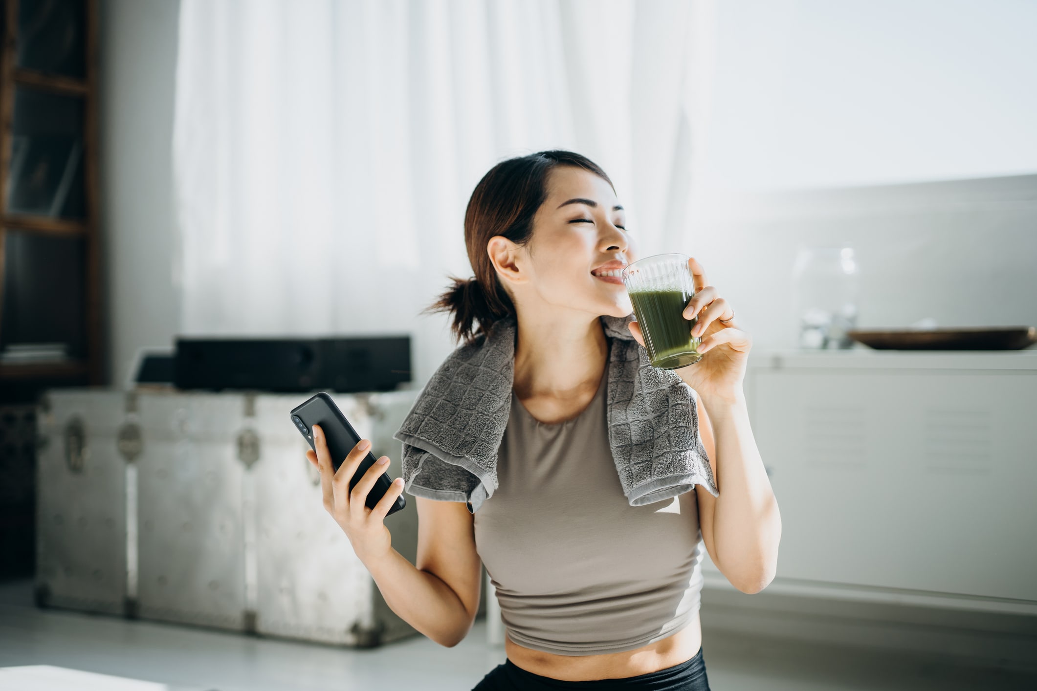 Young Asian Sports Woman Resting And Having Green Smoothie After Exercising At Home In The Morning, Using Smartphone