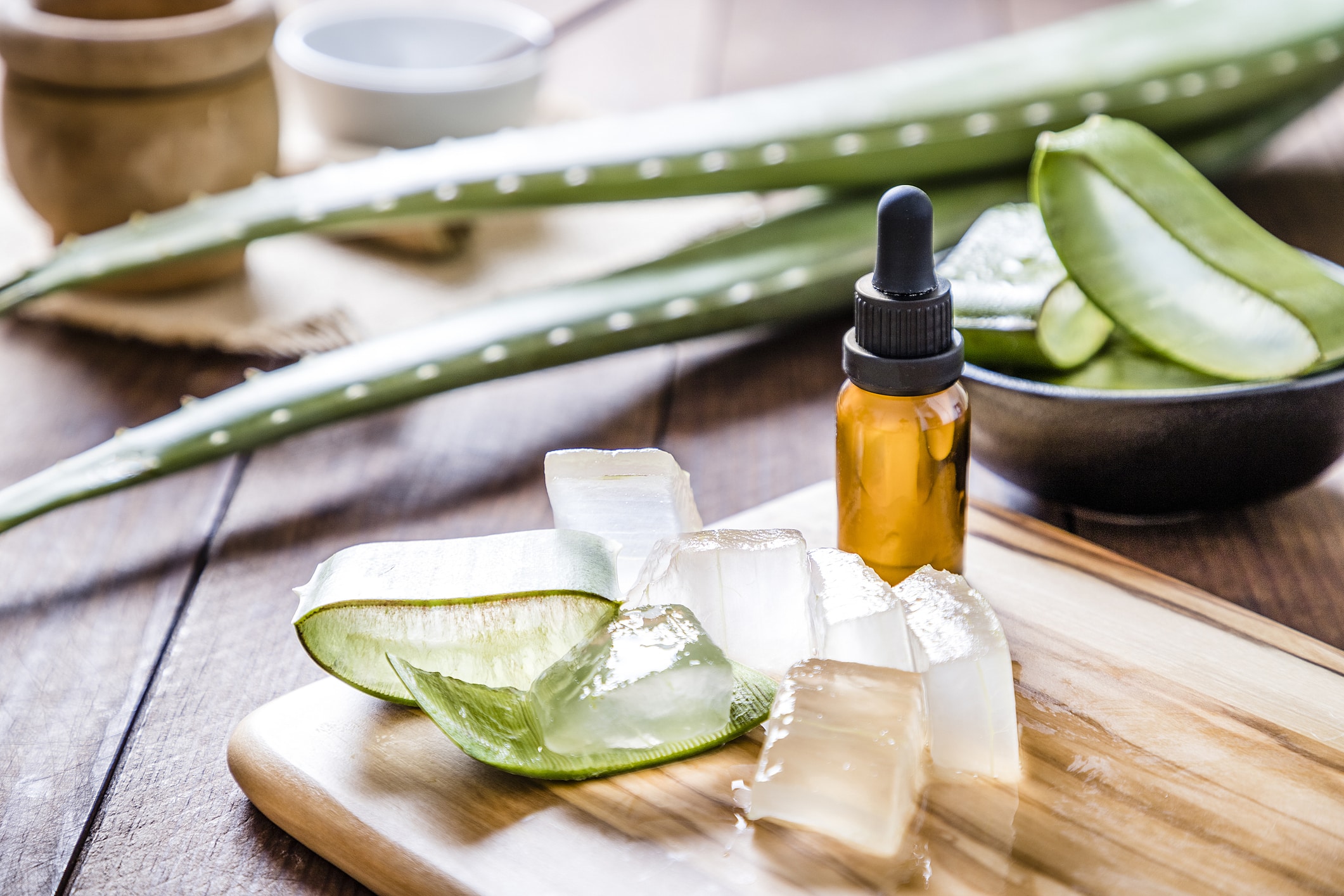 High Angle View Of Some Aloe Vera Crystals With A Cosmetic Dropper On A Wooden Board. A Black Bowl Full Of Sliced Aloe Vera Leaves Is Defocused On The Background.  Predominant Color Are Green And Brown. Studio Shot Taken With Canon Eos 6D Mark Ii And Canon Ef 24-105 Mm F/4L