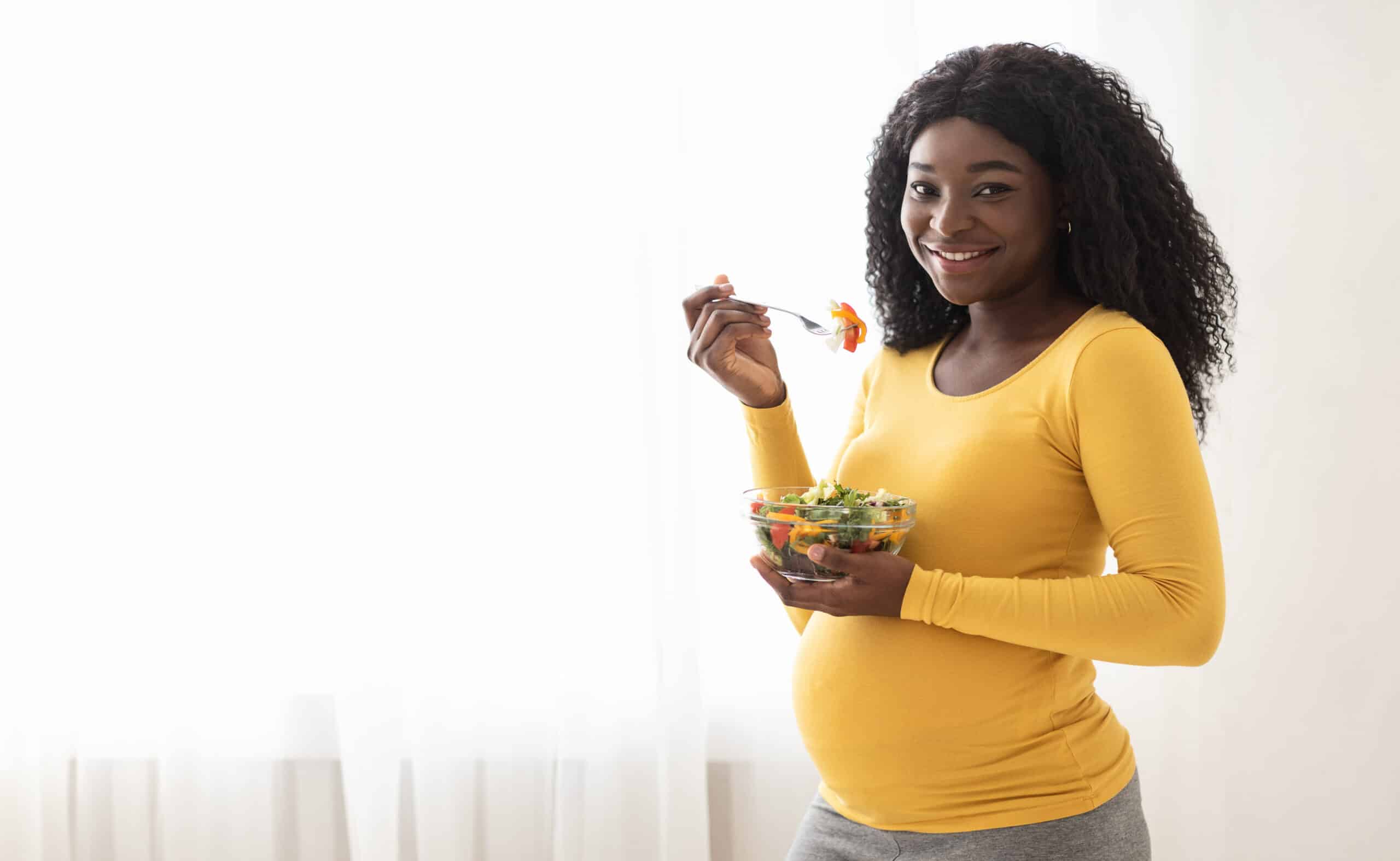 Nutrition And Healthy Diet During Pregnancy. Happy Pregnant Black Woman Enjoying Fresh Vegetable Salad At Home, Panorama With Copy Space. Cheerful Expecting African Lady Standing By Window And Eating