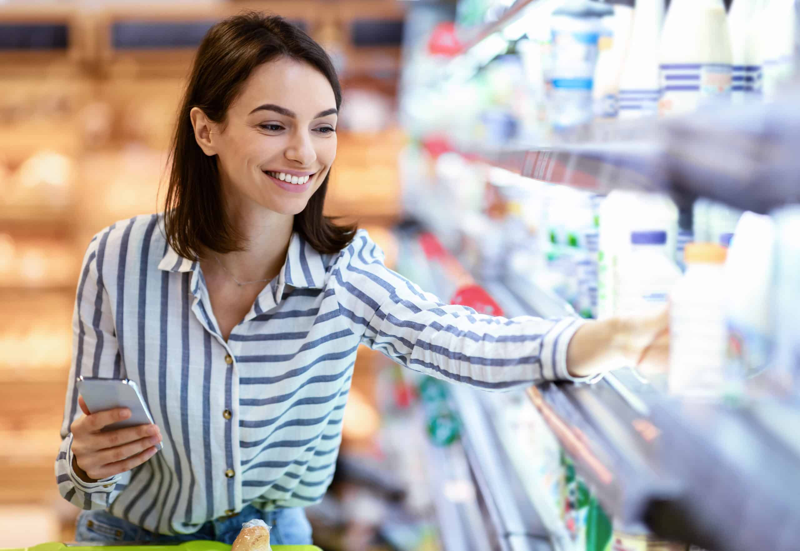 Shopping. Young Smiling Woman Holding And Using Phone Buying Food Groceries Standing In Supermarket. Female Customer With Smartphone Taking Milk From The Fridge In Dairy Section At The Shop