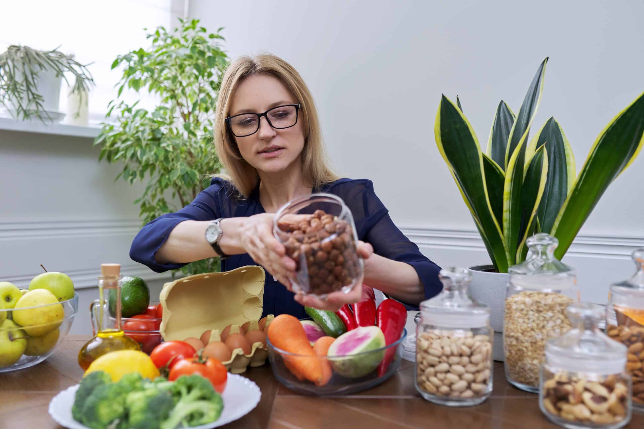 Middle Aged Woman Professional Nutritionist Sitting At Table With Vegetables Fruits Eggs Cereals Nuts, Looking At Camera, Doing Consultation, Holding In Hand Hazel Nuts