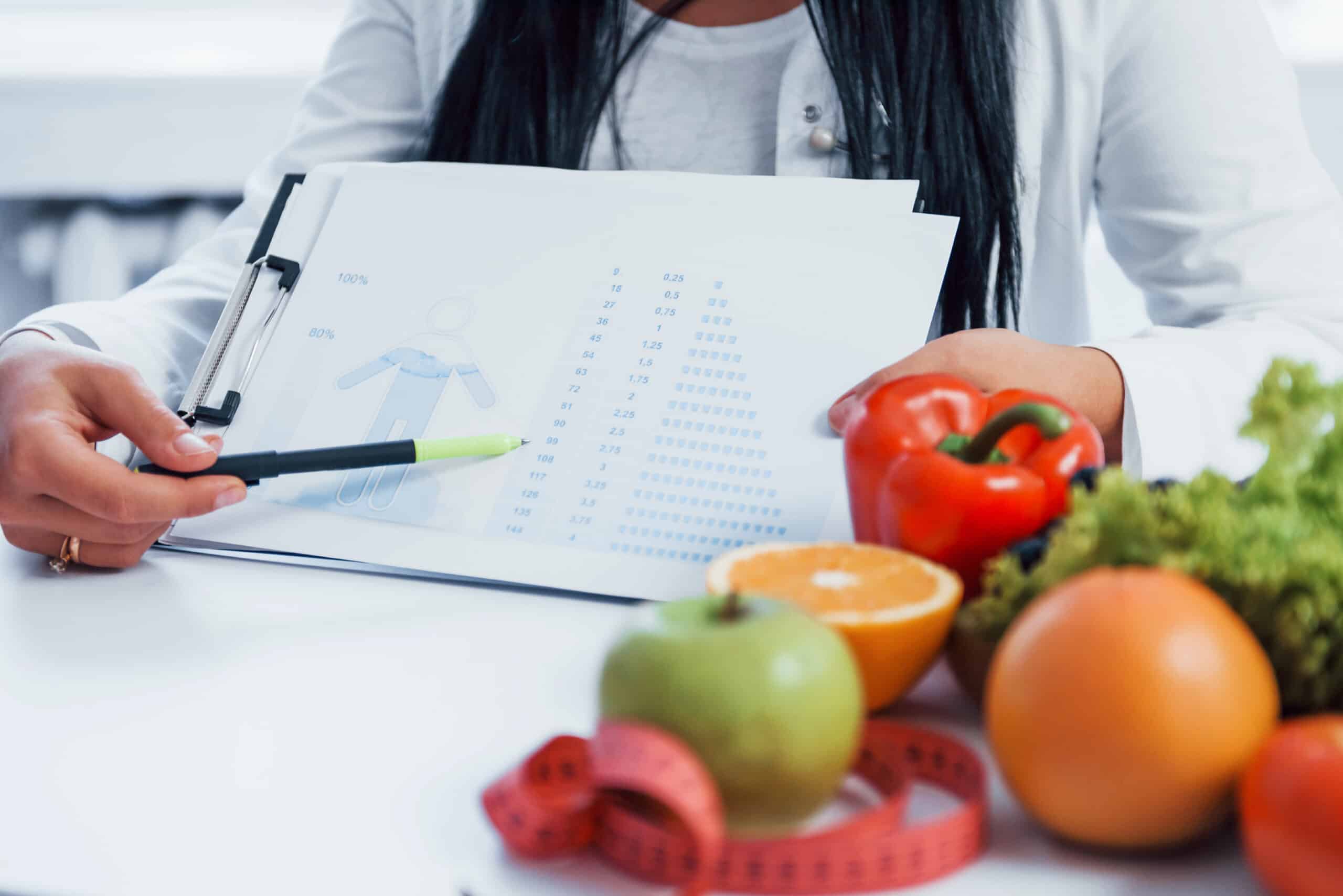 Female Nutritionist In White Coat Sitting Indoors In The Office At Workplace And Showing Information On Document.