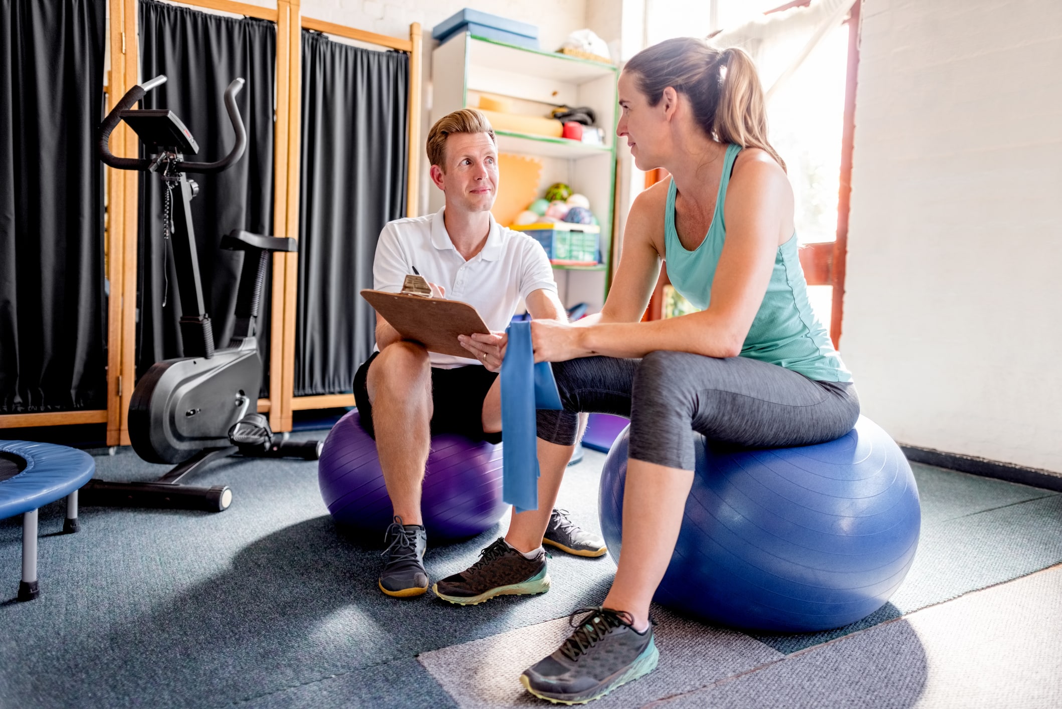 Woman And Her Personal Trainer Sitting On Fitness Balls And Discussing Exercising Plan At Gym