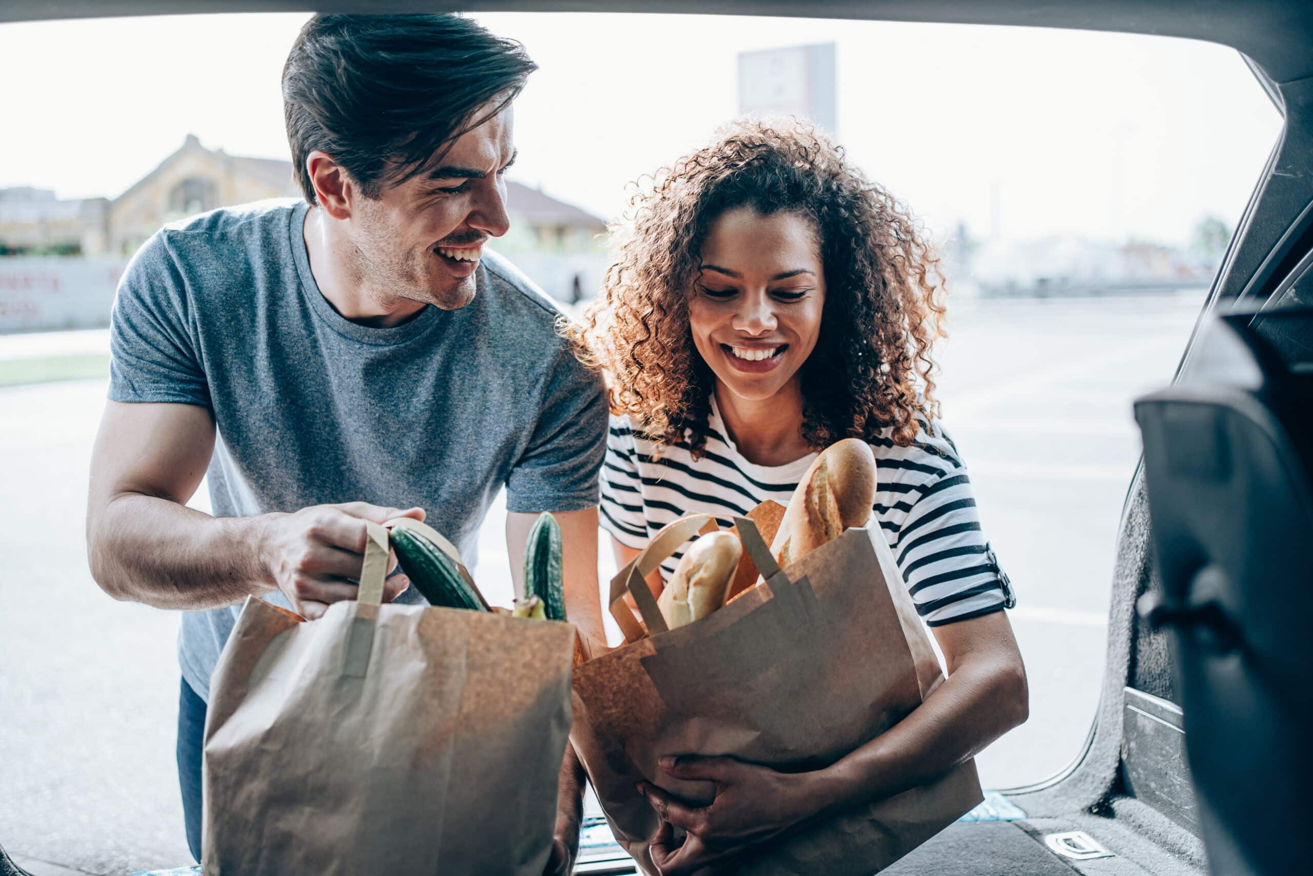 Shot Of An Adorable Smiling Multi-Ethnic Young Couple Loading Paper Shopping Bags In Their Car Trunk.