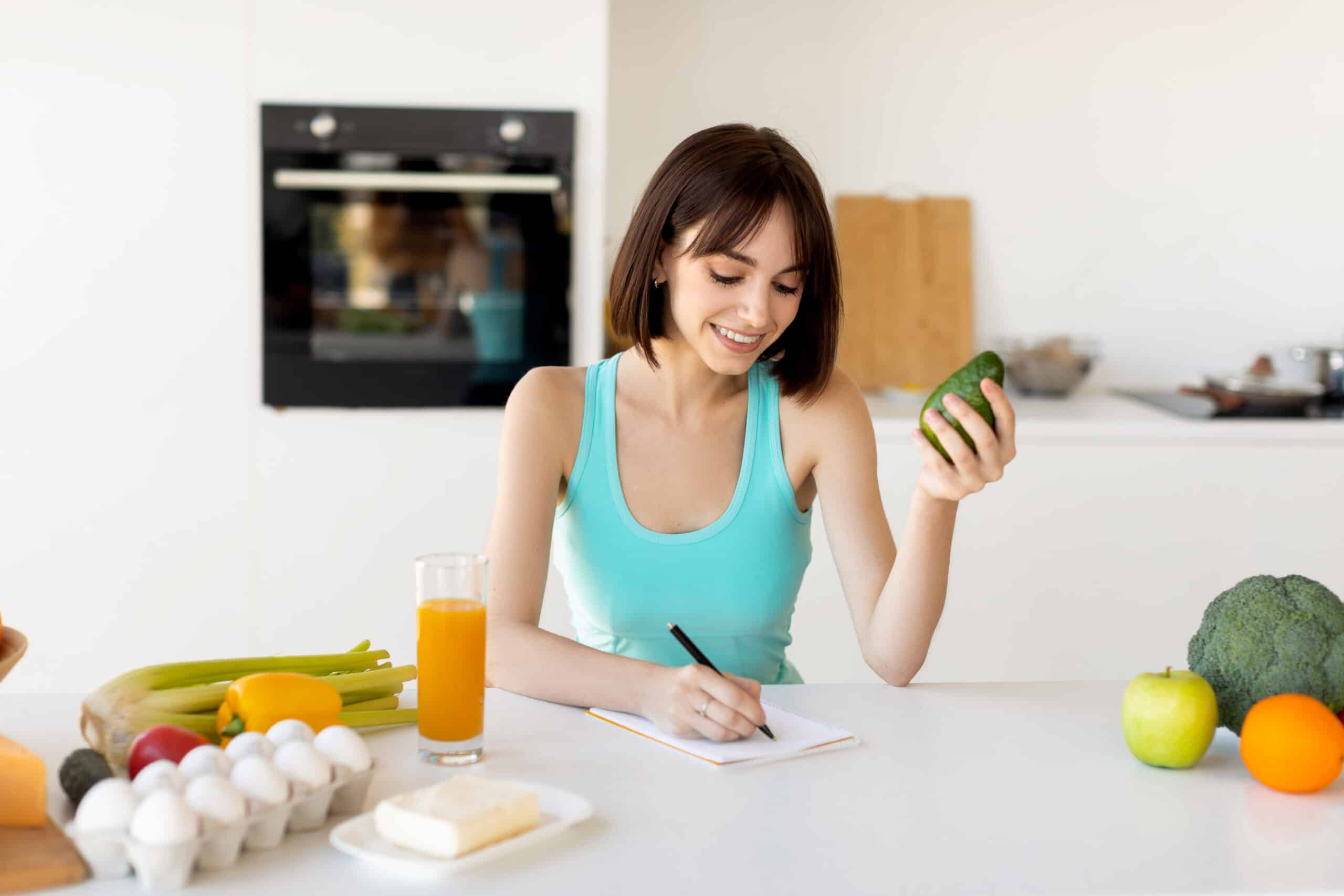 Keto Diet Concept. Happy Fit Woman Writing Daily Ration Diet Or Menu In Notebook, Sitting Near Various Products At Kitchen Table, Free Space
