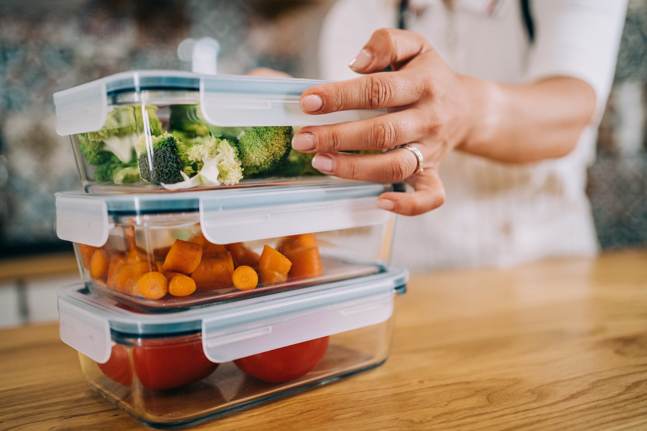 Hands Stacking Three Clear Glass Food Storage Containers With Snap-Lock Lids Containing Broccoli, Diced Carrots, And Tomatoes On Wooden Counter