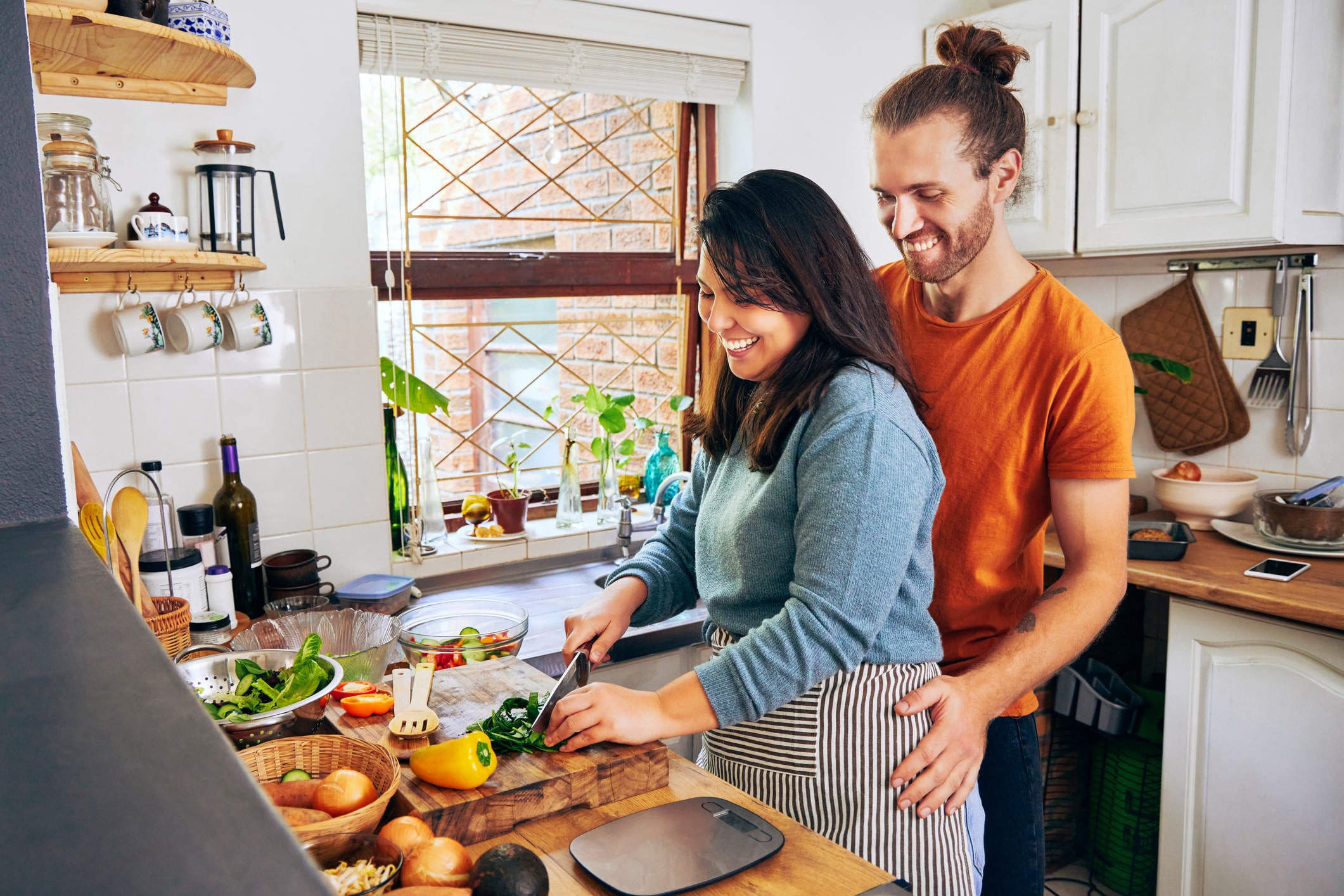 Couple Cooking Together In A Bright Kitchen, Woman In Teal Sweater Chopping Vegetables On Cutting Board While Man In Orange Shirt Stands Behind Her, Both Smiling