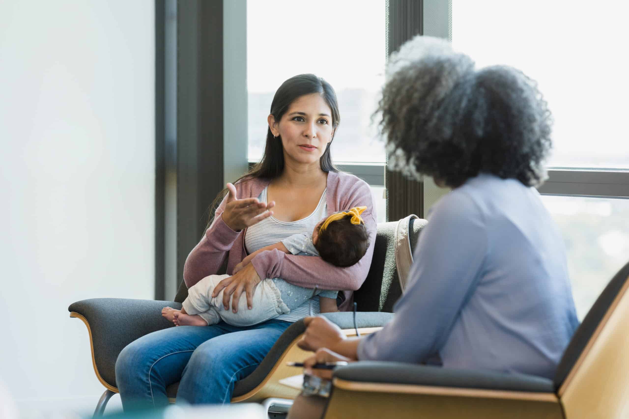 Postpartum Dietitian 1 Dietitian Group The Mid Adult Woman With A Baby In Her Arms Gestures As She Shares Her Struggles With Her Female Therapist.