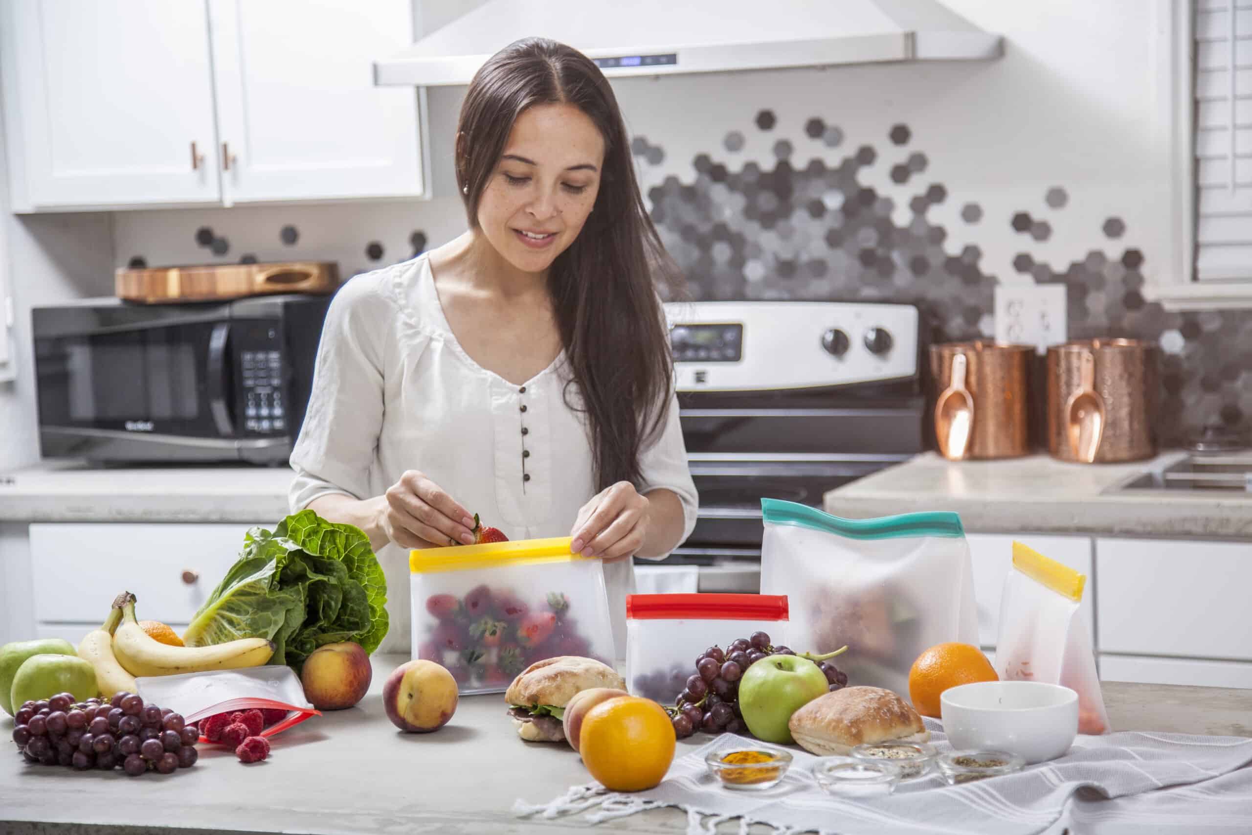 A Young Mother Puts Strawberries Into A Clear Food Storage Bag. She Is In The Kitchen With A Concrete Counter Top Surrounded By Other Fruits And Vegetables That She Is Preparing And Storing.