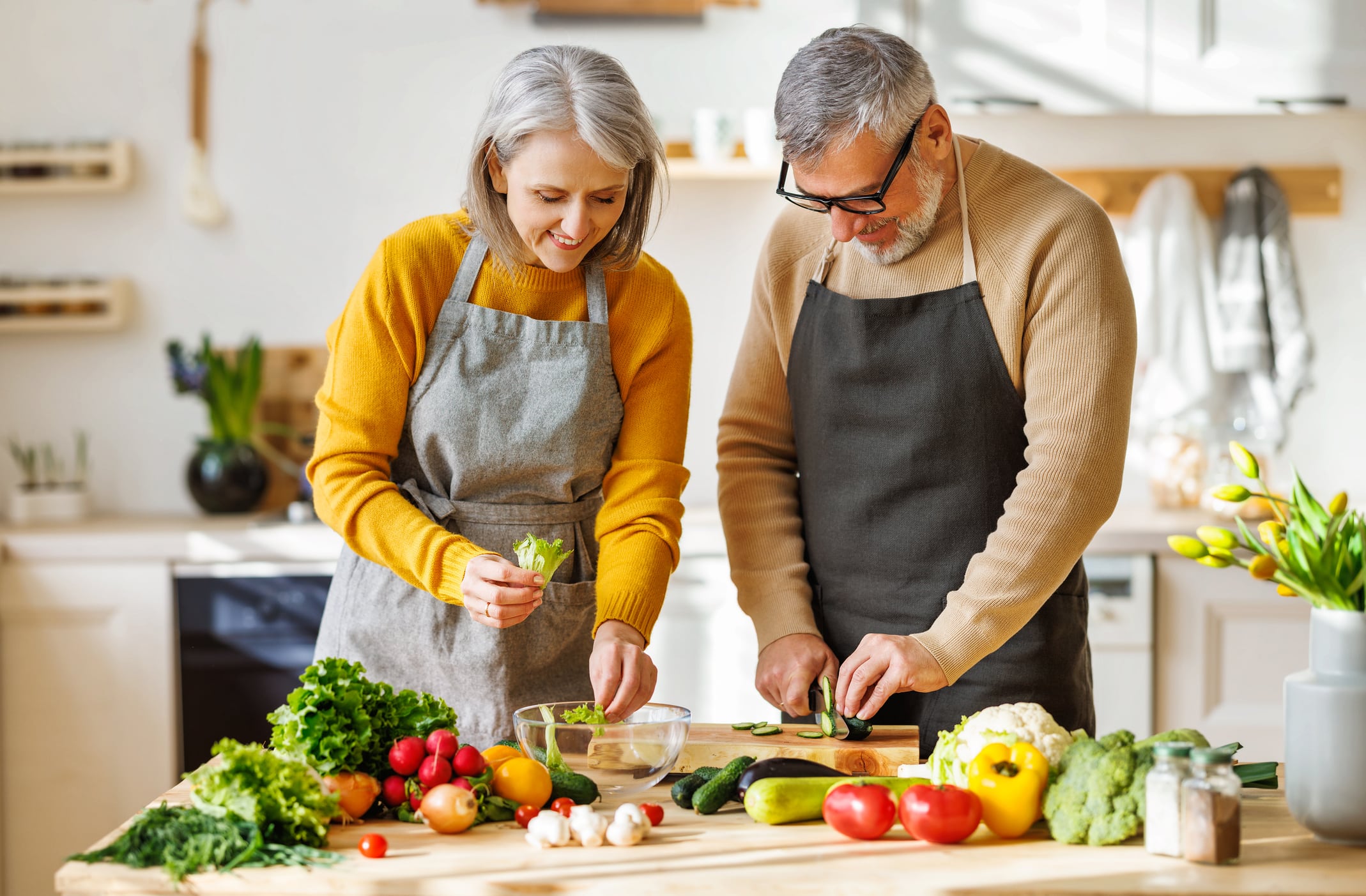 Happy Elderly Couple Smiling Husband And Wife In Aprons Prepare Salad Together At Kitchen Table, Chopping Variety Of Colorful Vegetables, Trying To Maintain Healthy Lifestyle Eating Vegetarian Food