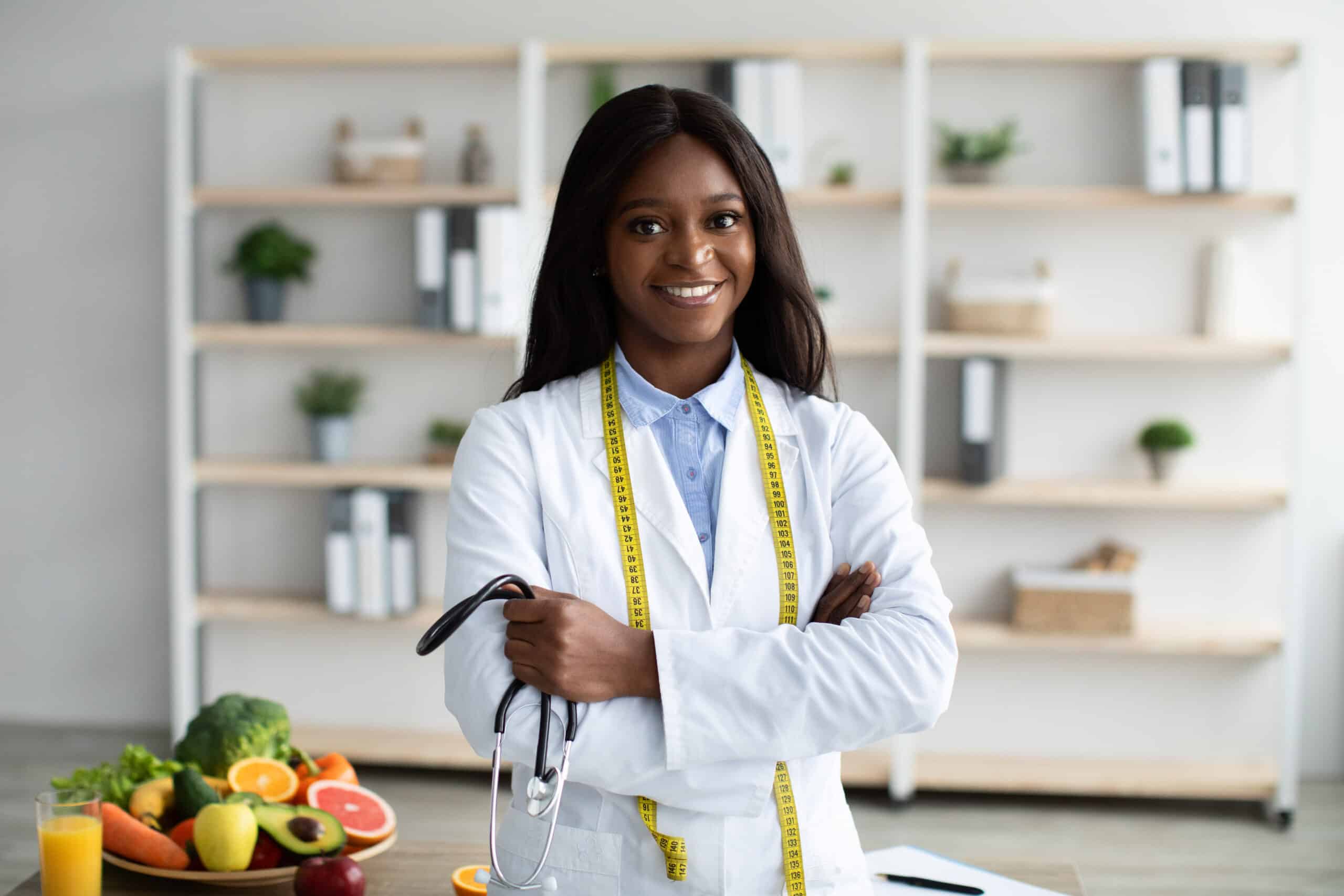 Portrait Of Friendly African American Nutritionist With Measuring Tape And Stethoscope In Hand Recommending Healthy Eating And Diet, Posing In Clinic Office