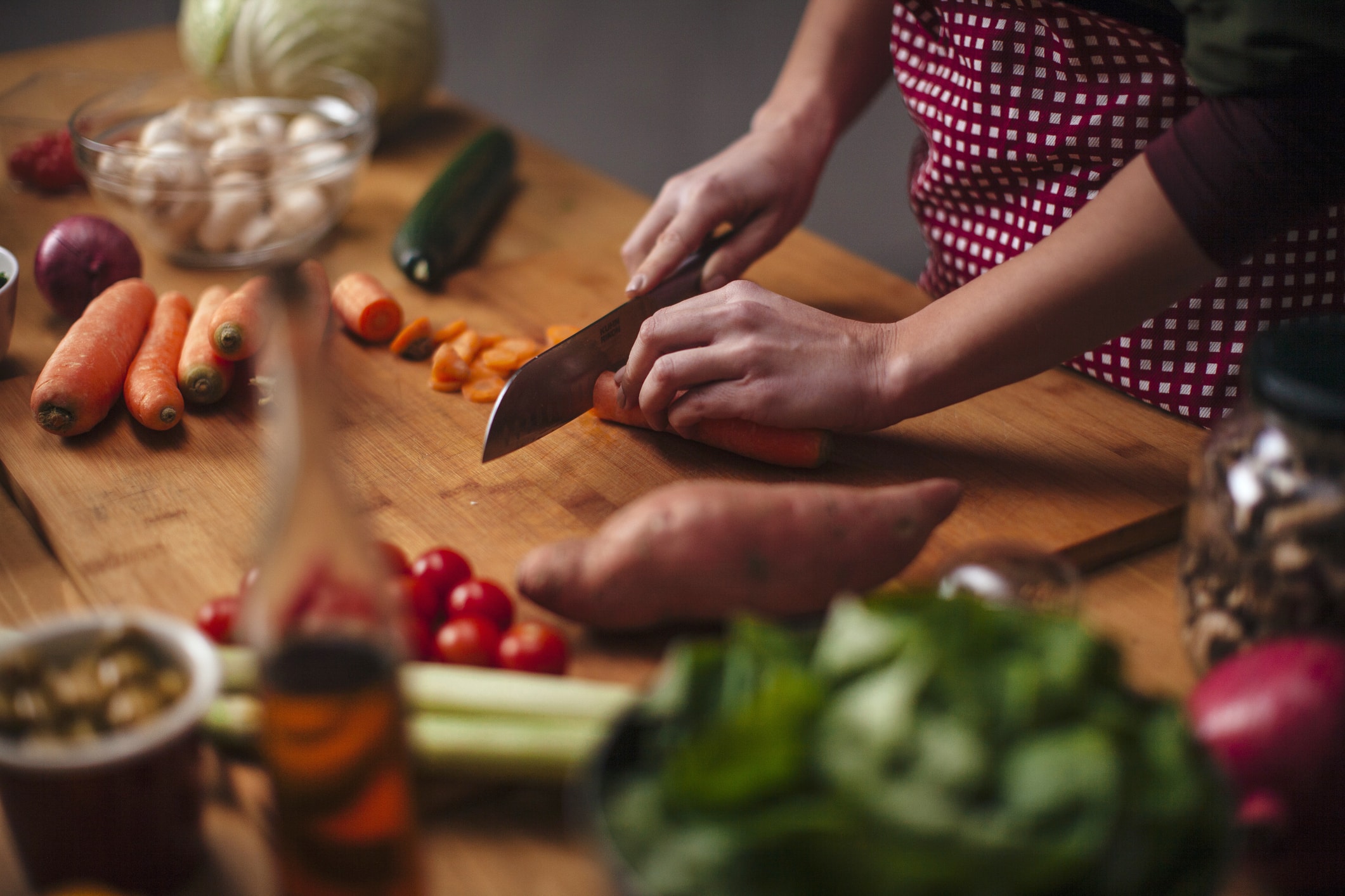 Hands Of Young Woman As She Chopping Carrots With Big Knife With Various Vegetables Around On Kitchen Counter