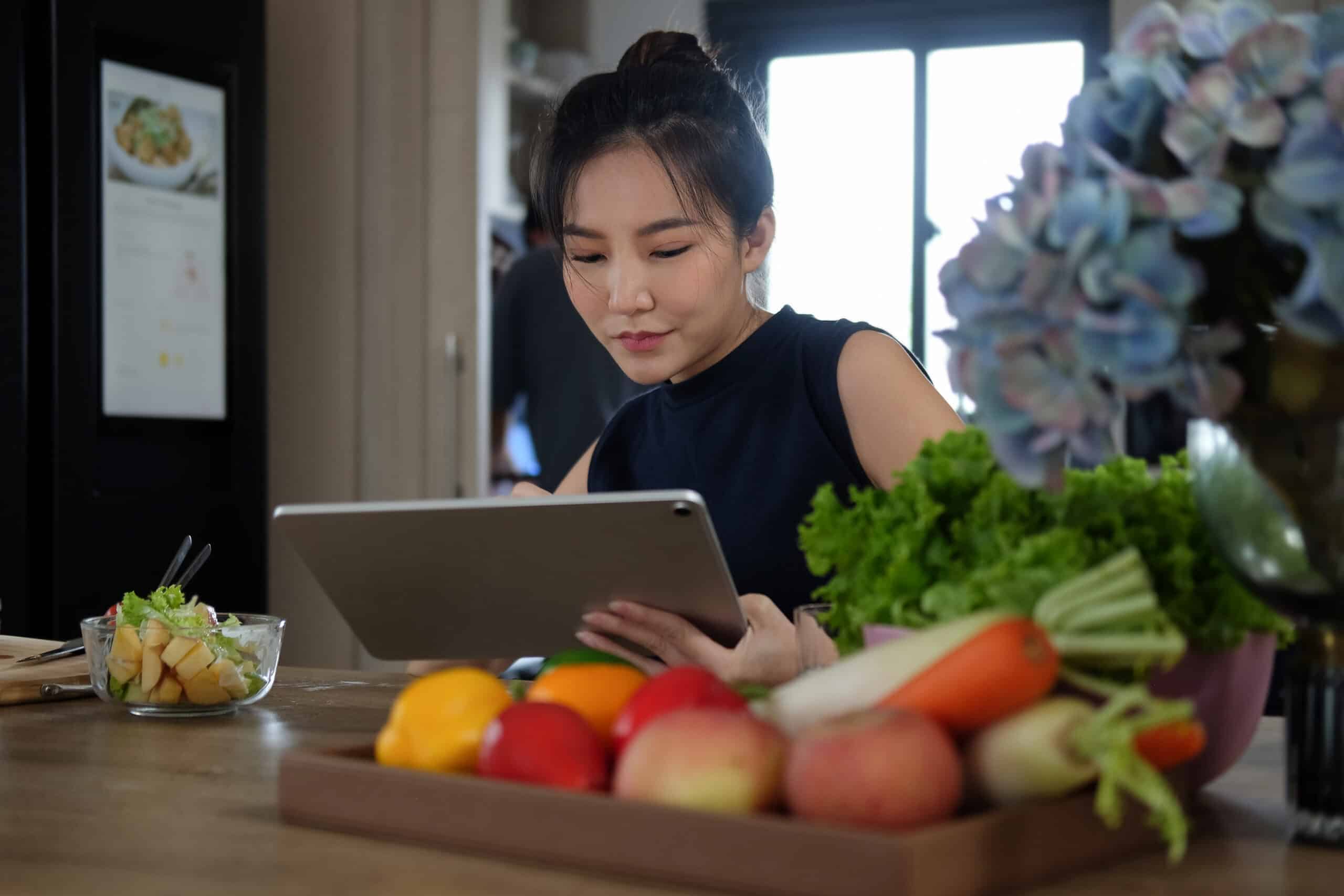 Woman Sitting At Kitchen Table Full Of Fresh Vegetables And Searching Recipes On Digital Tablet.