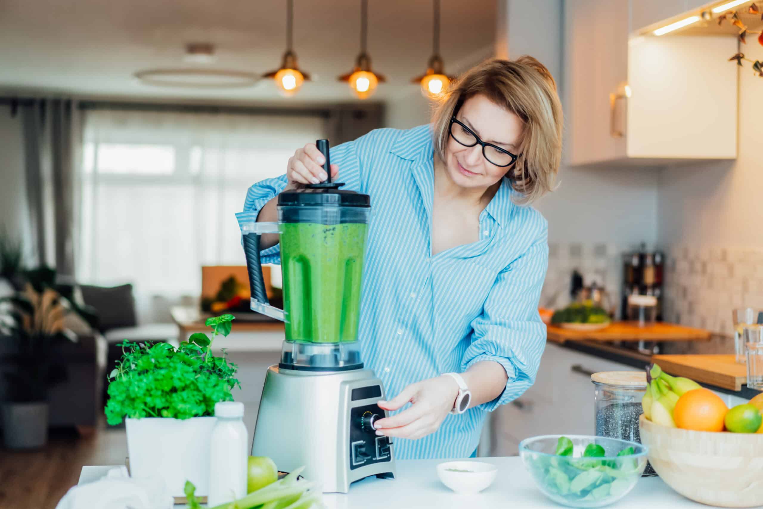 Middle Aged Woman With Blender And Green Vegetables Making Detox Shake Or Green Smoothie At Home. Healthy Dieting, Eating, Cooking. Natural Anti Aging, Weight Loss Program. Vegan, Vegetarian Diet.