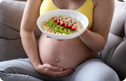 Expecting parent holds their pregnant belly while holding a bowl of oatmeal with slices of colored fruit.