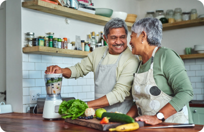 Adding fruit and vegetables to a blender, a couple smiles at each other in their kitchen.