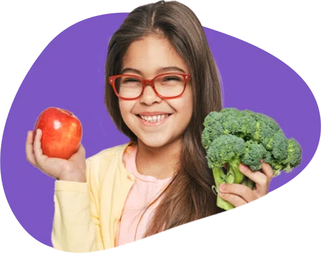 A kid smiling at the camera while holding broccoli and apple