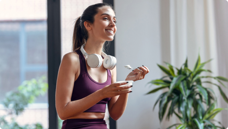 A lady in an exercising outfit and headphones around her neck, eating yogurt