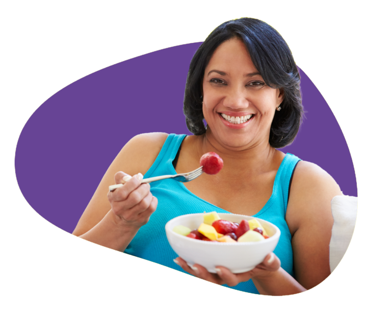 Smiling at the camera, person holds a white bowl in one hand filled with various fruits, and holds a fork with a strawberry in the other hand.