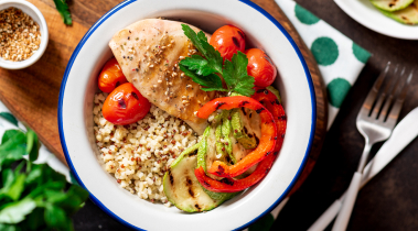 White and blue bowl sit on a table with brown rice, grilled vegetables and chicken inside it.
