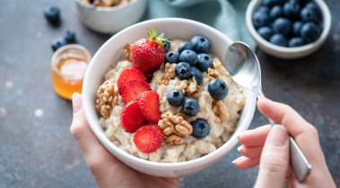 Hands hold a white bowl of oatmeal with fresh strawberries and blueberries.