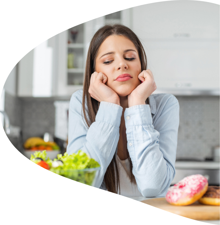 A person holding their face in their hands looks contemplatively at a bowl of salad and a couple of donuts in front of them.