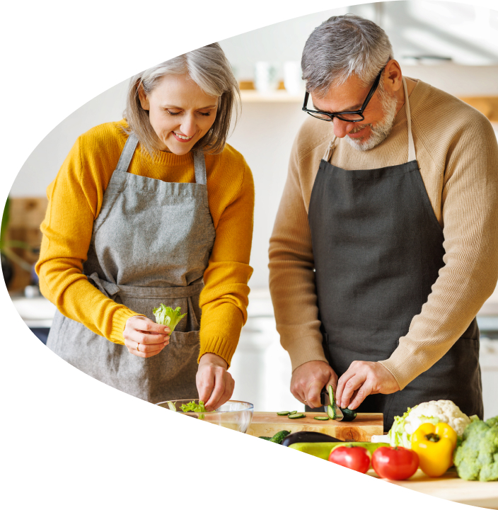 A couple stand inside their kitchen cutting vegetables on a cutting board, wearing aprons and smiling.