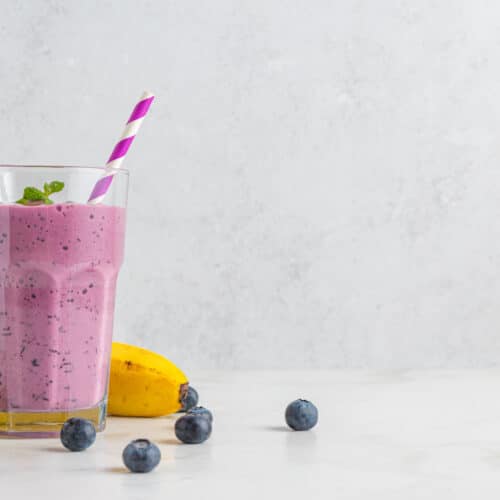 Blueberry and banana smoothie or milkshake in a glass with straw, fresh berries and mint on white background. Summer refreshing drink