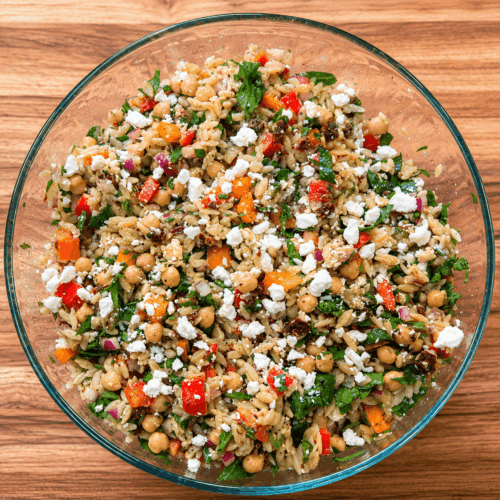Chickpea Orzo Salad With Feta, Sun-Dried Tomatoes, Spinach, And Pine Nuts In A Glass Bowl