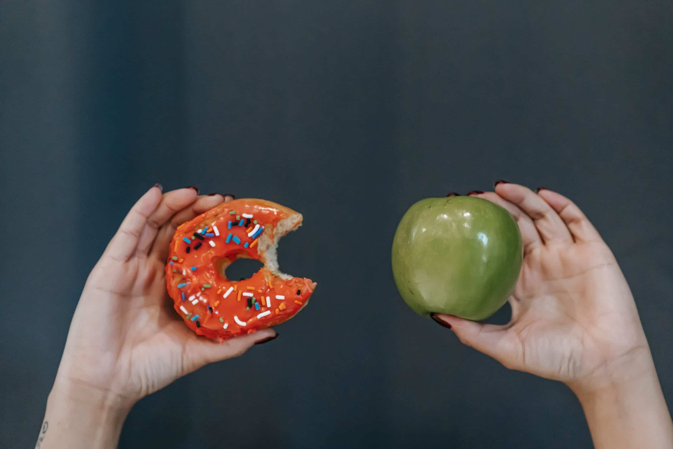 Hands Holding A Partially Eaten Sprinkled Donut In One Hand And A Green Apple In The Other Against A Dark Background.